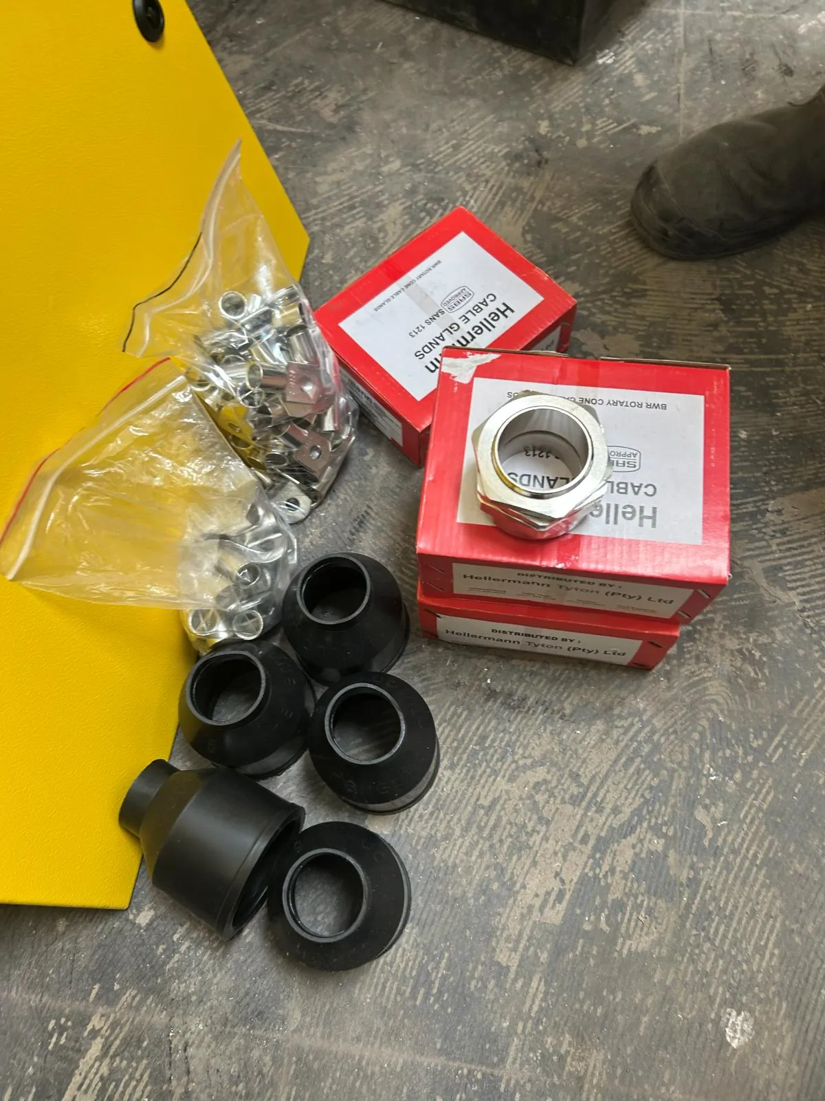 A brass armoured cable gland on its box beside black rubber shrouds and a bag of cable cleats