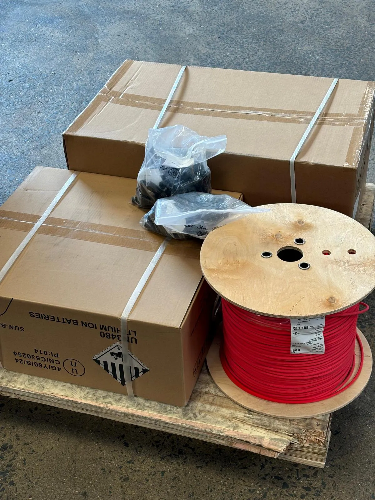 Cartons of lithium-ion batteries on a pallet beside bagged connectors and a drum of red solar cable