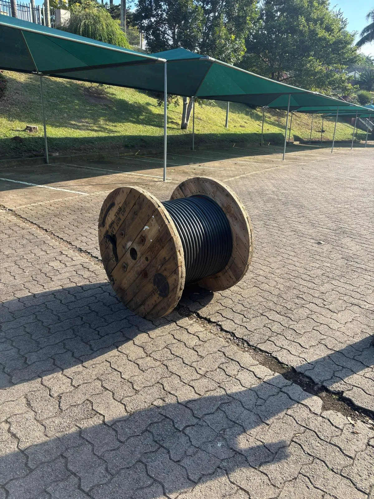 A wooden drum of black cable standing on paving under shade netting.