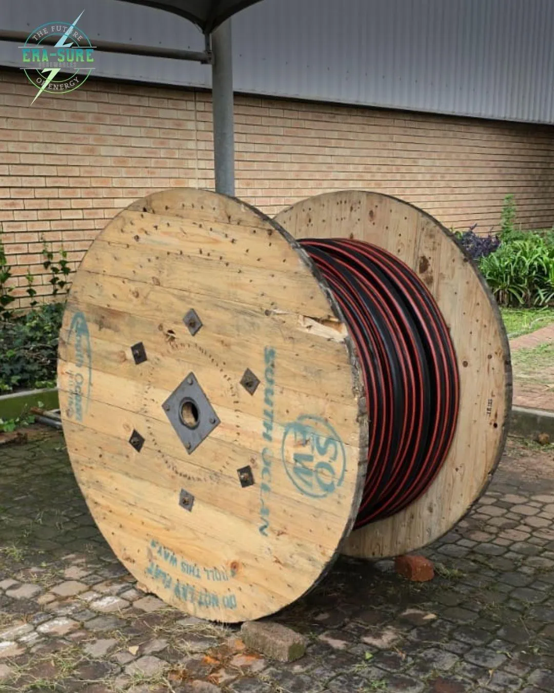 A wooden drum of red and black cable standing beside a brick building.