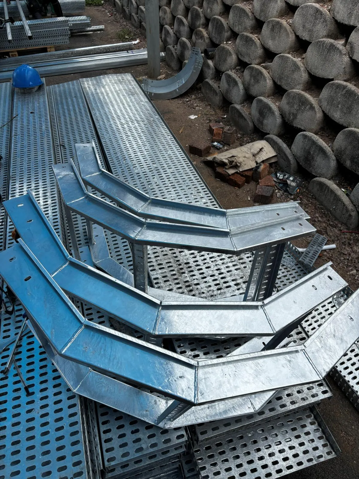 Galvanised cable-tray bends and risers stacked on top of straight perforated tray lengths in a yard.