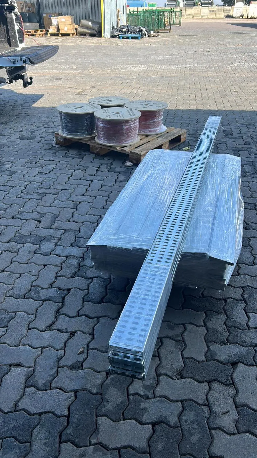 A wrapped pallet of cable tray with a single perforated tray length resting on it, beside a pallet of cable drums.