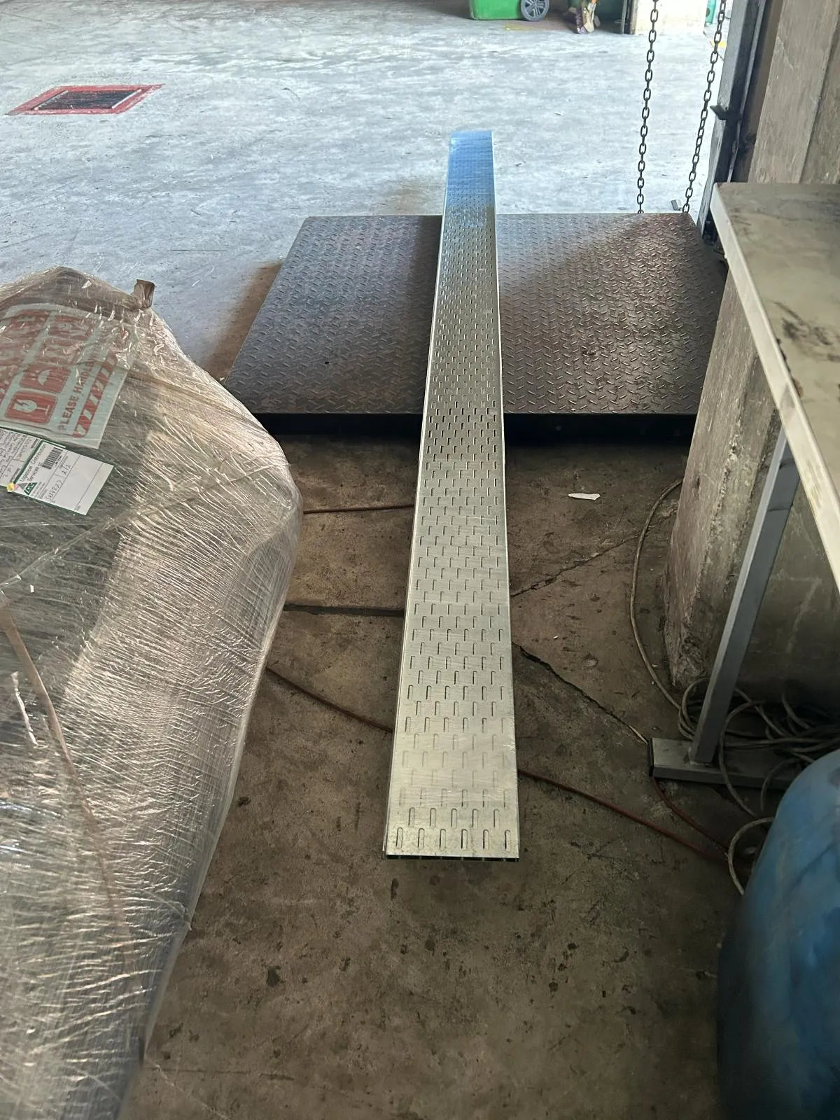 A narrow perforated cable tray laid across a workshop floor beside a wrapped pallet.