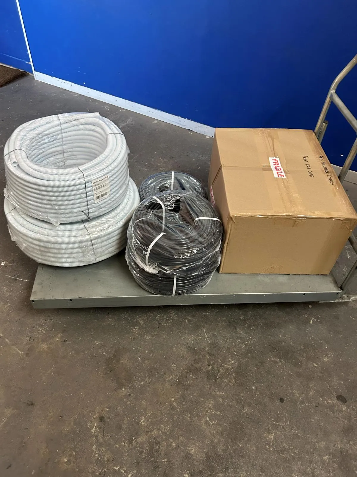 Coils of white and black flexible conduit beside a carton marked fragile on a warehouse trolley