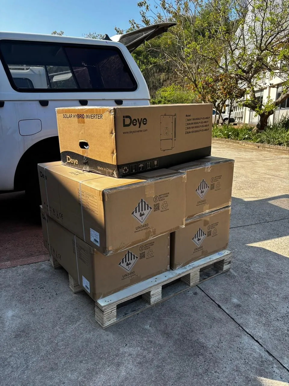 A boxed Deye hybrid inverter stacked above four battery cartons on a pallet beside a delivery vehicle