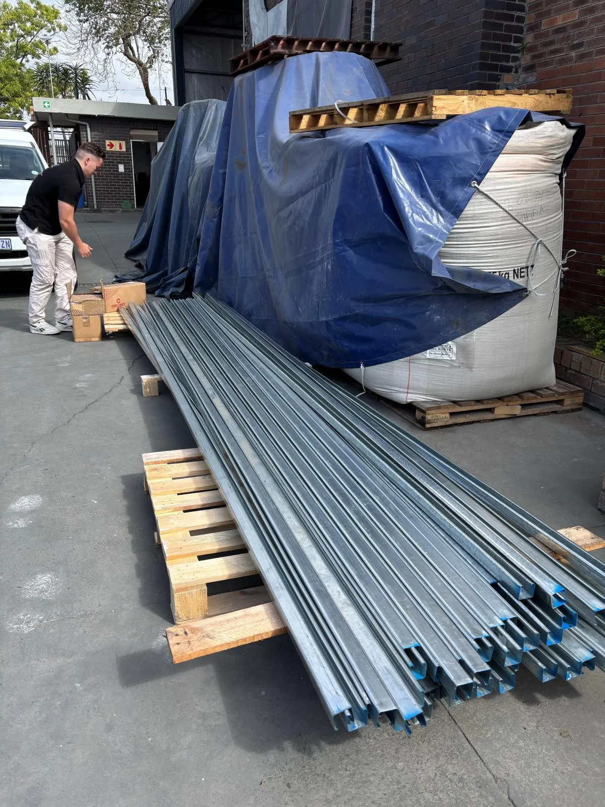 Long lengths of galvanised channel and support strut stacked on pallets in a yard, with an Era-Sure team member checking the delivery against the paperwork.