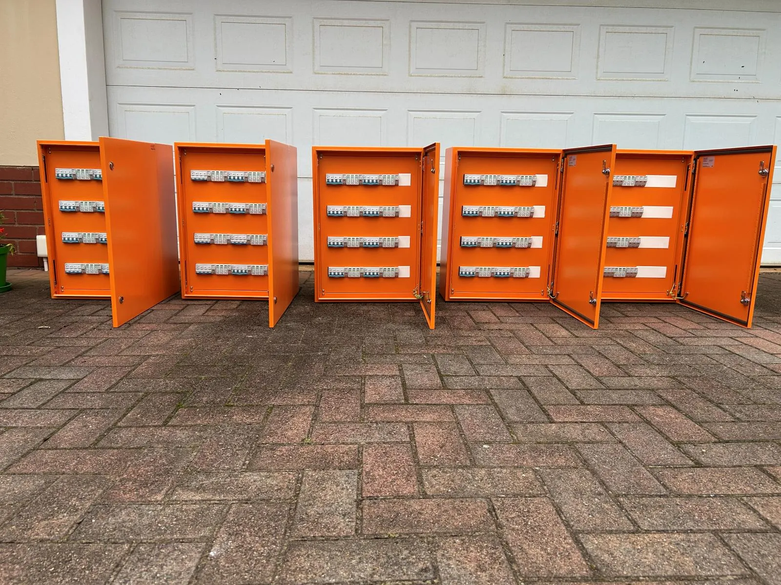 Six orange steel enclosures standing open on paving, each populated with rows of breakers.