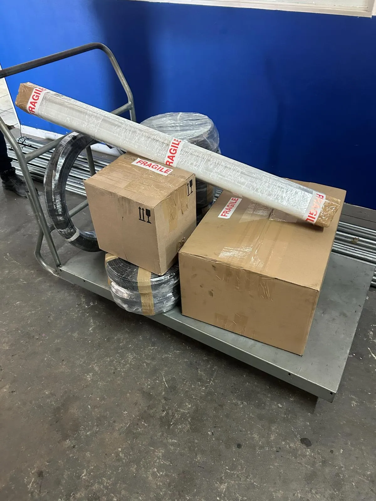Cartons marked fragile, coiled conduit and a wrapped length of trunking stacked on a warehouse trolley